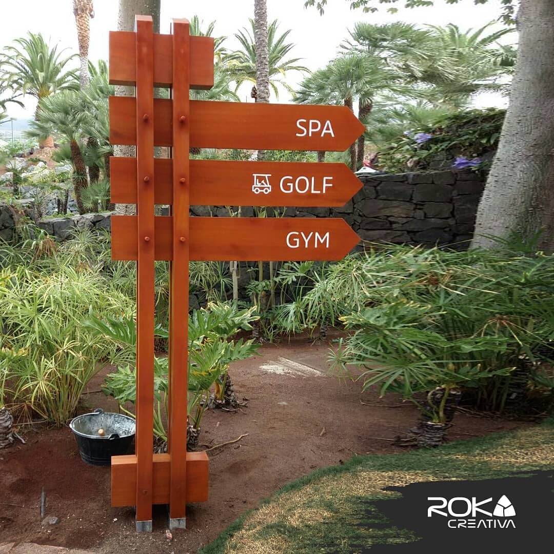 Poste de señalética en madera con flechas hacia Spa, Golf y Gym entre la vegetación